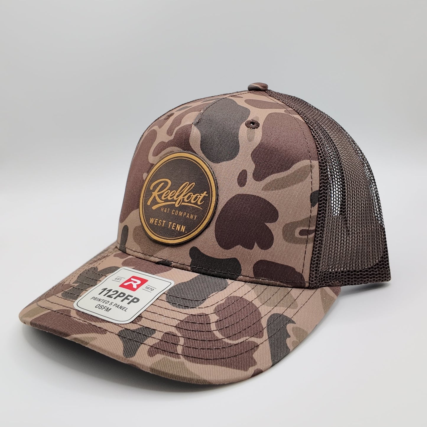 A camouflage trucker hat with a brown mesh back, featuring a circular 'Reelfoot hat co" patch on the front and a richardson brand sticker on the brim, set against a neutral gray background.