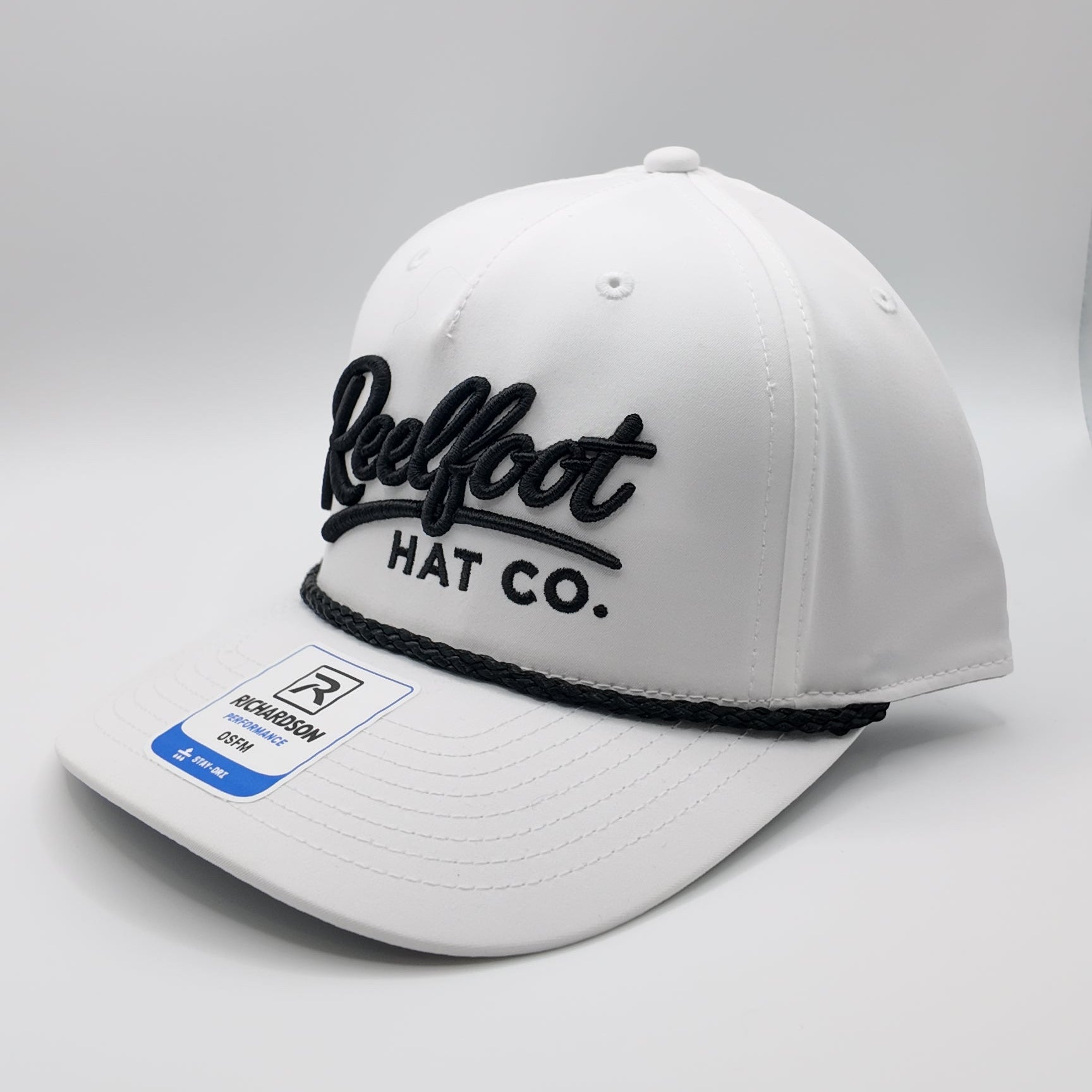 A white structured baseball cap with black embroidered 'Reelfoot HAT CO.' lettering on the front and a black elastic band under the brim, shown on a light gray studio background.