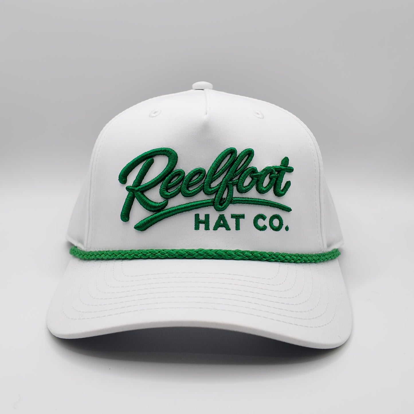 A white structured baseball cap with green embroidered 'Reelfoot HAT CO.' lettering on the front and a green braided rope detail across the brim, shown on a light gray studio background.