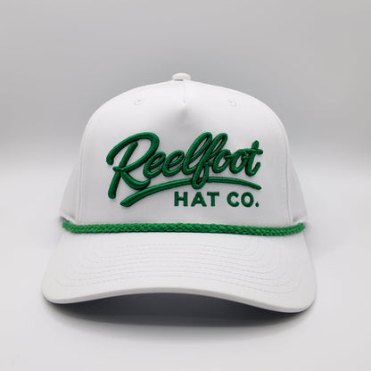 A white structured baseball cap with green embroidered 'Reelfoot HAT CO.' lettering on the front and a green braided rope detail across the brim, shown on a light gray studio background.