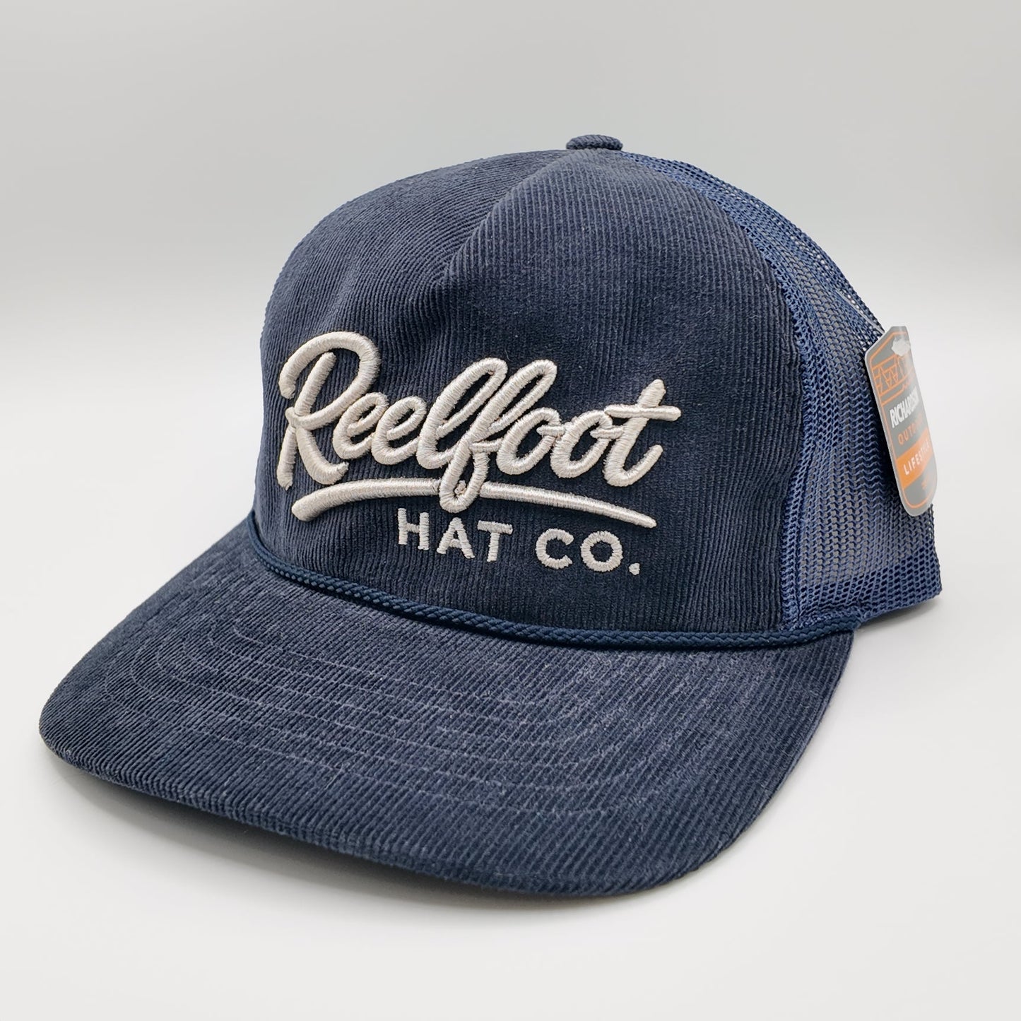 A navy blue trucker hat with a washed denim-like front panel and mesh back, featuring 'Reelfoot HAT CO.' embroidered in white on the front.