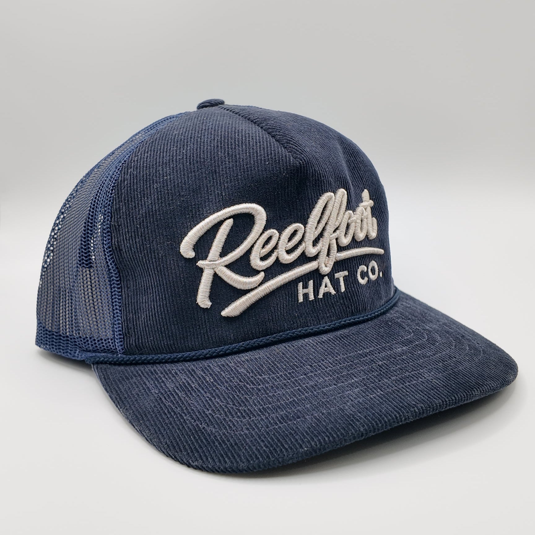 A navy blue trucker hat with a textured front panel and mesh back, featuring 'Reelfoots HAT CO.' embroidered in white script on the front.
