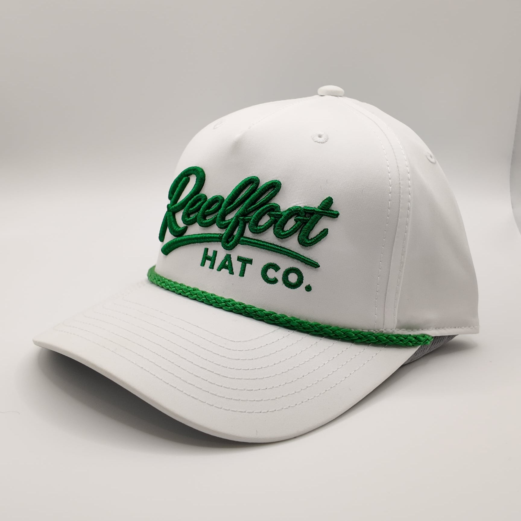 A white baseball cap with green embroidered text reading 'Reelfoot HAT CO.' on the front and a green braided trim along the brim, photographed on a light gray studio background.