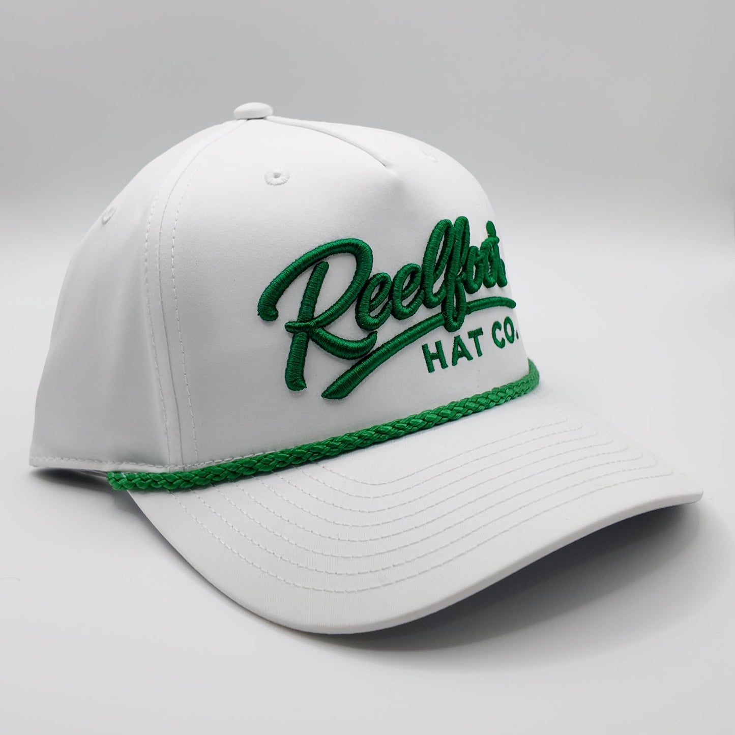 A white baseball cap with green embroidered 'ReelLot HAT CO' text on the front and a green braided trim along the brim, shown on a light gray studio background.