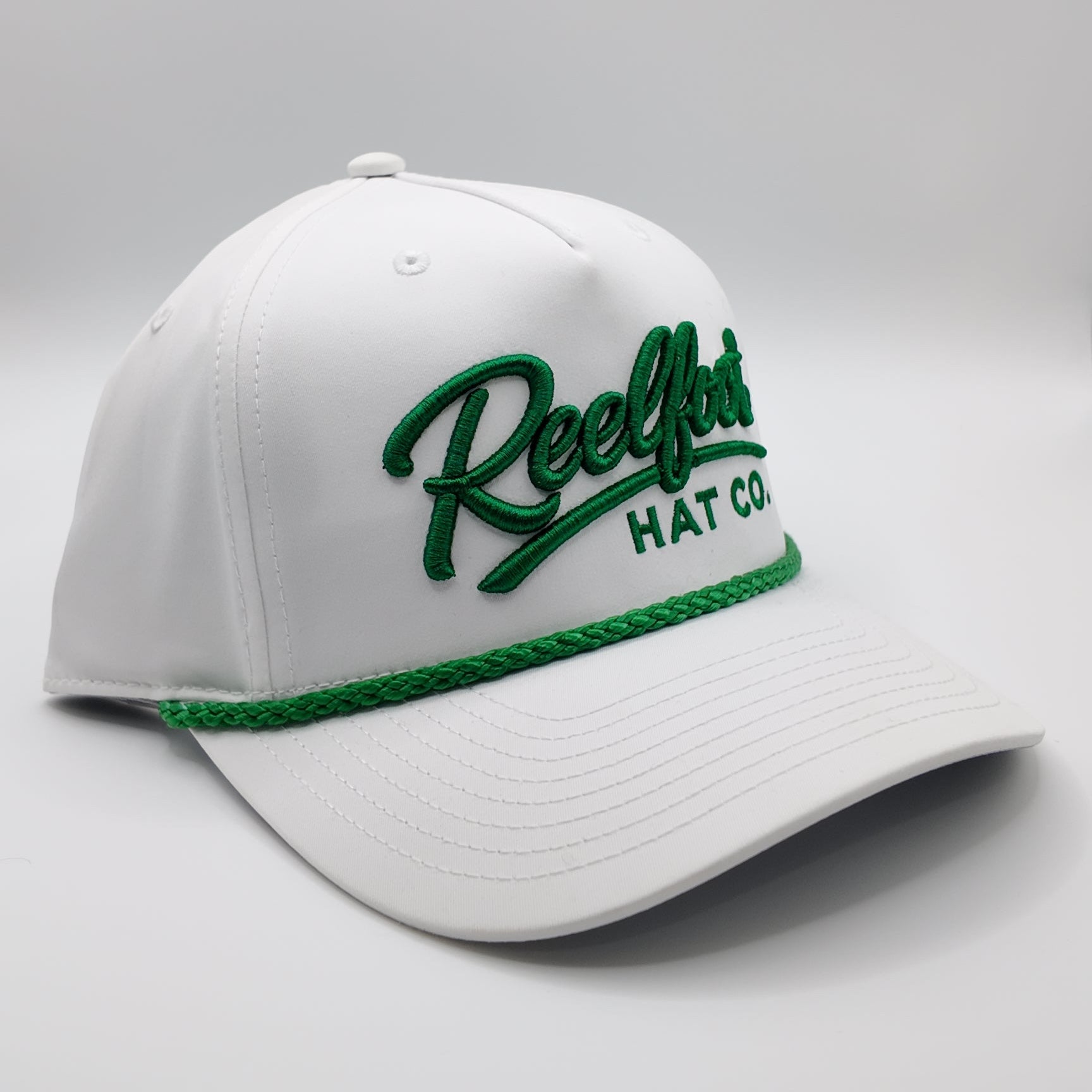 A white baseball cap with green embroidered 'ReelLot HAT CO' text on the front and a green braided trim along the brim, shown on a light gray studio background.